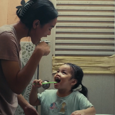 A woman and young child brushing their teeth together in a small bathroom, smiling at each other.
