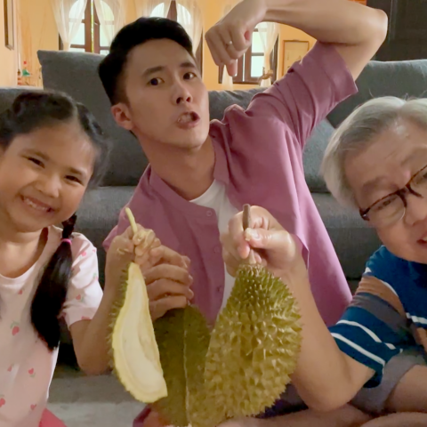 Multigenerational family sitting on a living room floor smiling while opening and holding a durian fruit together.