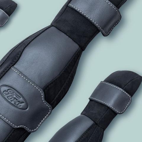 Three Ford SupportBelt™ padded seatbelt covers displayed on a sage green background, showing the black and grey faux leather construction, white contrast stitching, adjustable straps, and embossed Ford logo.