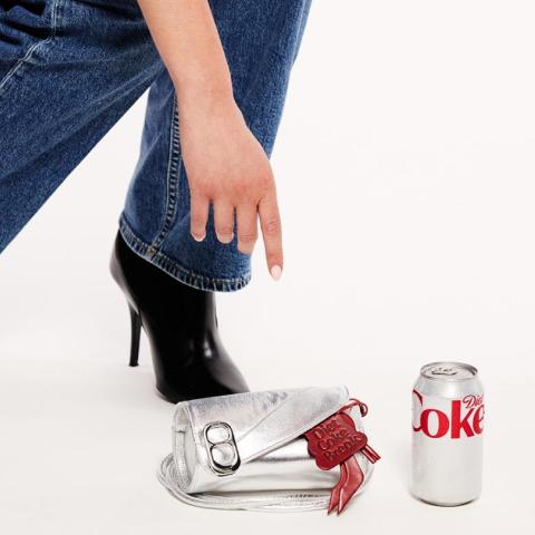 A silver metallic clutch bag designed to look like a crushed Diet Coke can, featuring a pull-tab detail and red bow tag reading 'Diet Coke Break', placed next to a real Diet Coke can on a white surface, with a model in wide-leg blue jeans and black stiletto ankle boots reaching toward it