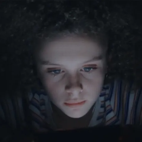 A girl with blonde curly hair is watching a film at night. The scene is so dark that the screen is lighting up her face.