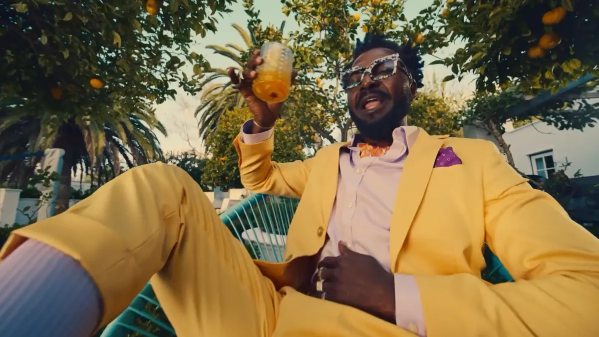 Person wearing a yellow suit sits outdoors in a garden holding a glass of orange drink, surrounded by citrus trees.