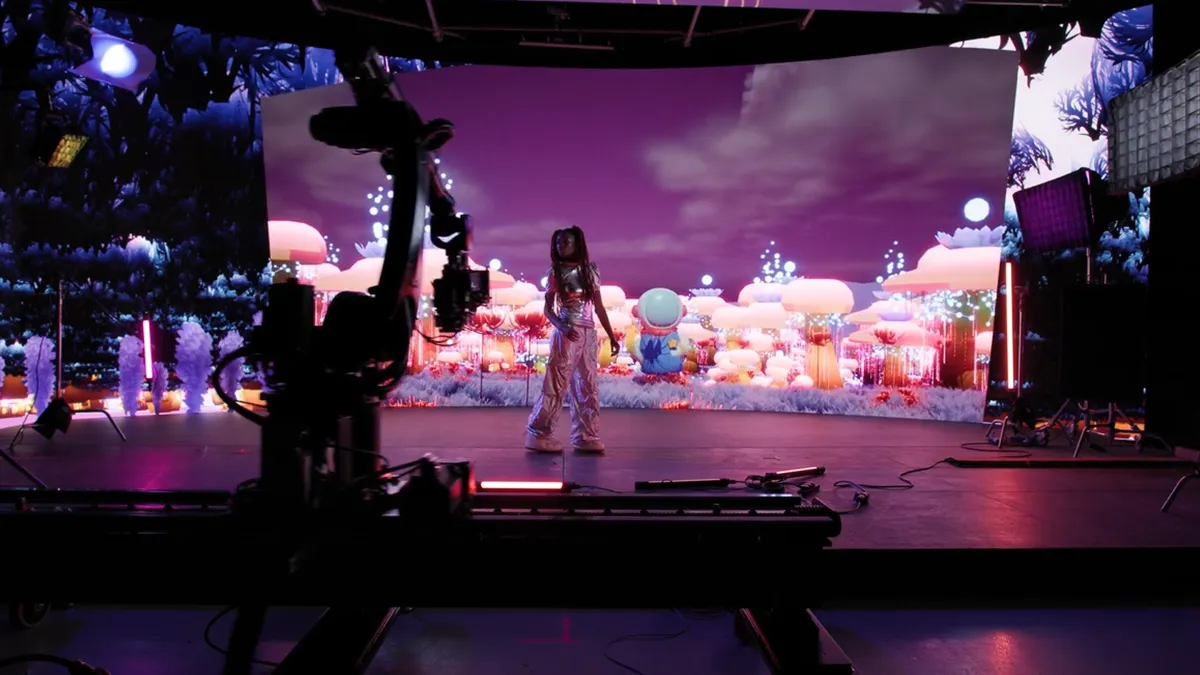 Wide shot of a virtual production stage with LED wall backdrop showing a colourful fantasy landscape and a performer standing centre stage.